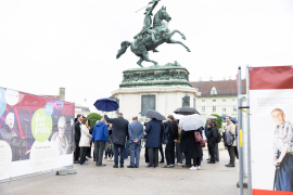 Blick auf eine Gruppe von Menschen mit Regenschirmen bei einer Pressekonferenz vor dem Erzherzog-Karl-Reiterdenkmal am Heldenplatz, flankiert von den Wänden der Gedenkausstellung am Heldenplatz © MKÖ/Alissar Najjar 