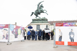 Blick auf eine Gruppe von Menschen mit Regenschirmen bei einer Pressekonferenz vor dem Erzherzog-Karl-Reiterdenkmal am Heldenplatz, flankiert von den Wänden der Gedenkausstellung am Heldenplatz © MKÖ/Alissar Najjar 