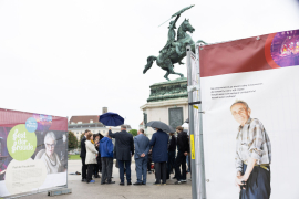 Blick auf eine Gruppe von Menschen mit Regenschirmen bei einer Pressekonferenz vor dem Erzherzog-Karl-Reiterdenkmal am Heldenplatz, flankiert von den Wänden der Gedenkausstellung am Heldenplatz © MKÖ/Alissar Najjar 